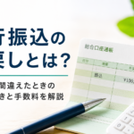 銀行振込の組戻し手続きを解説する記事の画像。通帳と電卓、ボールペンが置かれ、警告アイコンとともに「銀行振込の組戻しとは？ 振込先を間違えたときの返金手続きと手数料を解説」の文字が表示されている。